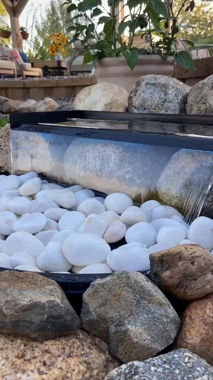 The finished spillway sends a curtain of water onto white pebbles inside a natural rock border.